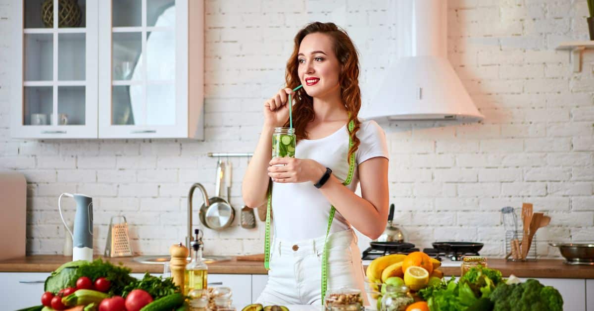 Young woman drinking fresh water with cucumber, lemon and leaves of mint from glass in the kitchen