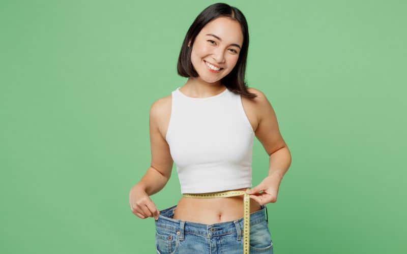 Young woman wear white clothes show loose pants after weightloss hold measure tape on waist isolated on plain pastel light green background