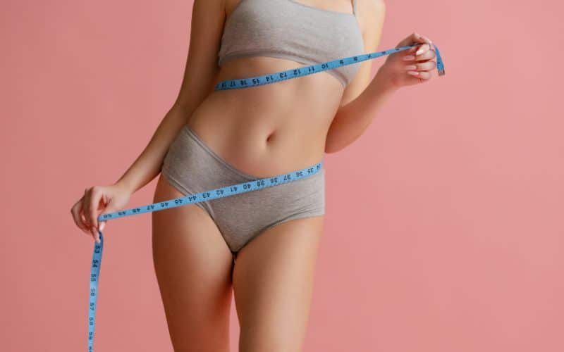  natural female body in gray inner wear, measuring waist isolated over pink background