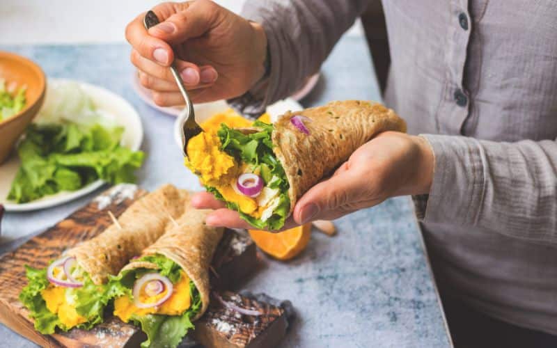  Woman make Mexican tortilla flatbread wraps stuffed with green salad leaves, onion and chickpea hummus filling in hands